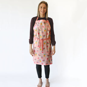 a smiling lady with blonde hair standing wearing a Bibetta Wipeezee adult apron in pink yummy treats pattern. She has fastened the waist straps at the front of the apron . She is wearing a black top and leggings and no shoes or socks.