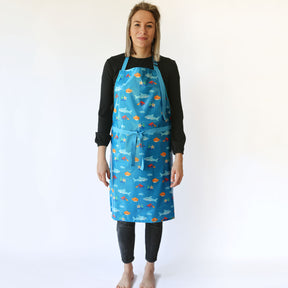 a smiling lady with blonde hair standing wearing a Bibetta Wipeezee adult apron in turquoise sea creatures pattern. She has fastened the waist straps at the front of the apron . She is wearing a black top and leggings and no shoes or socks.