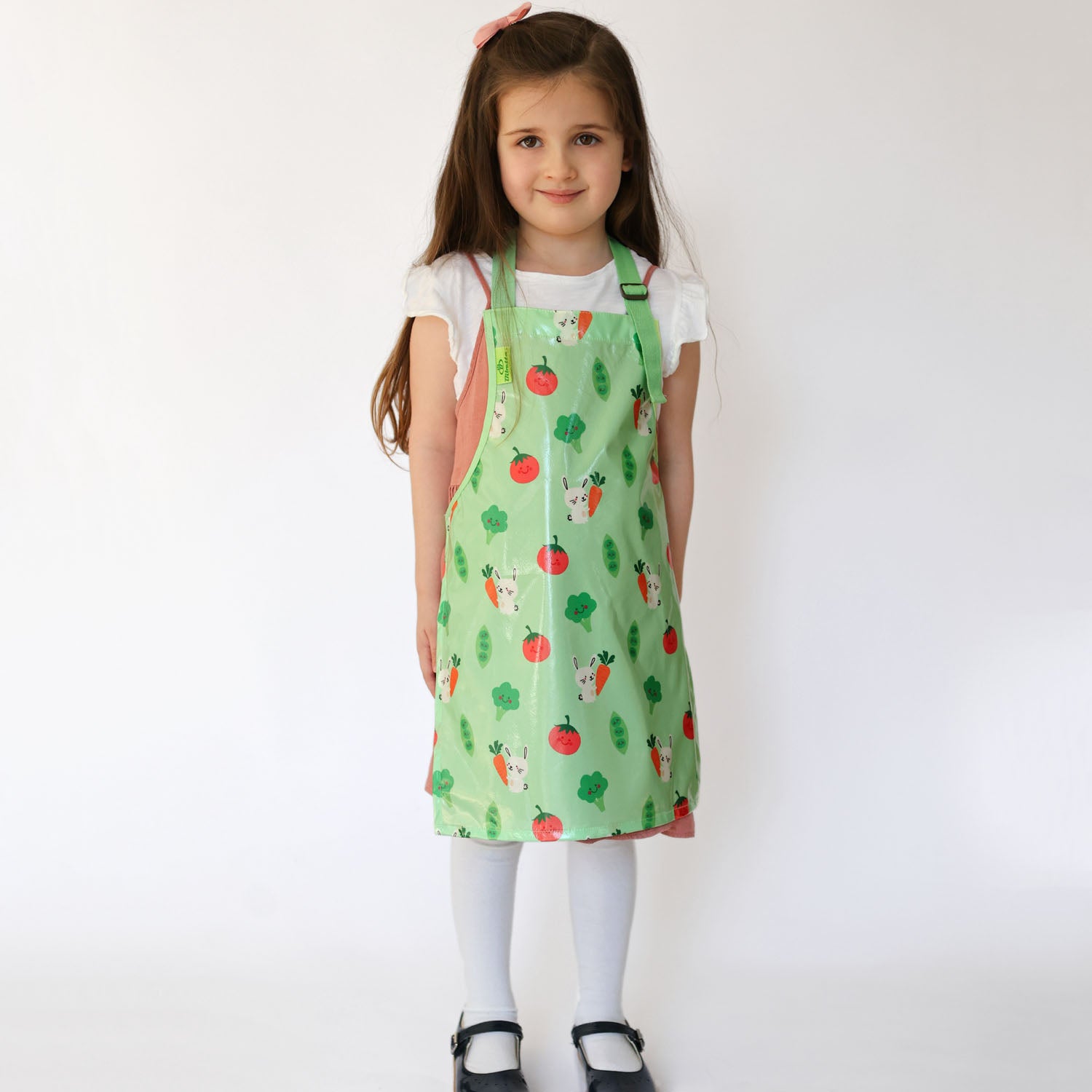 a smiling girl standing wearing a Bibetta Wipeezee childs apron in green bunny and veg pattern.  She is wearing a white t-shirt and pink pinafore with white tights and black shoes.