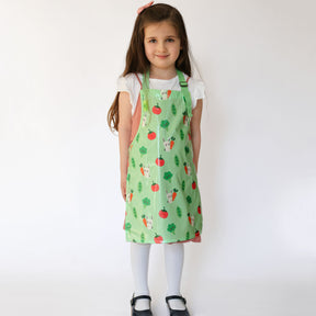 a smiling girl standing wearing a Bibetta Wipeezee childs apron in green bunny and veg pattern.  She is wearing a white t-shirt and pink pinafore with white tights and black shoes.