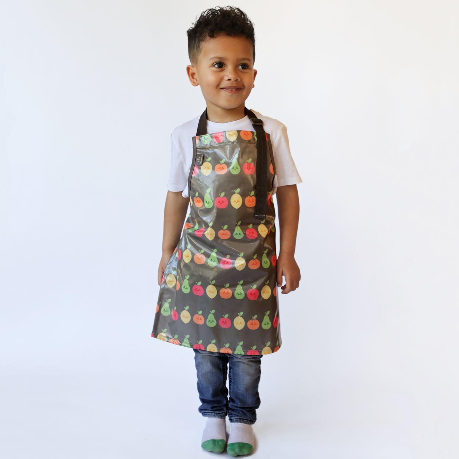 a smiling boy standing wearing a Bibetta Wipeezee childs apron in grey happy fruit pattern.  He is wearing a white t-shirt and blue jeans with white and green socks.