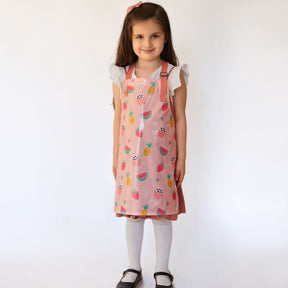 a smiling girl standing wearing a Bibetta Wipeezee childs apron in pink yummy treats pattern.  She is wearing a white t-shirt and pink pinafore with white tights and black shoes.