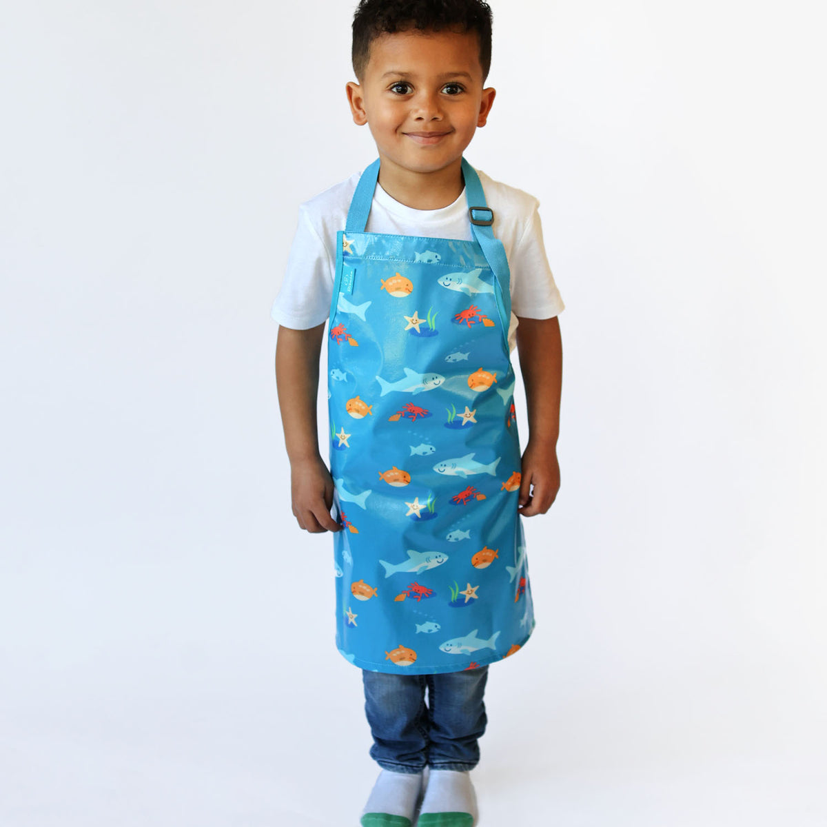 a smiling boy standing wearing a Bibetta Wipeezee childs apron in turquoise sea creatures pattern.  He is wearing a white t-shirt and blue jeans with white and green socks.