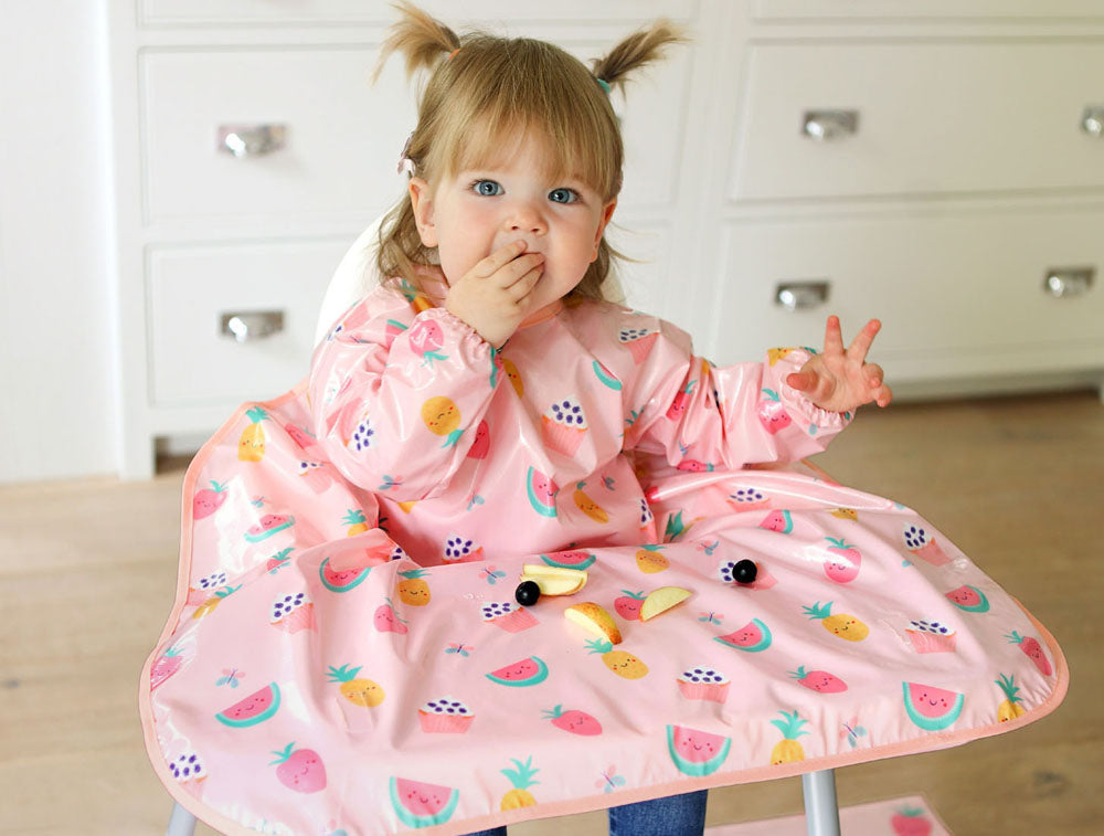 A happy toddler child sat in a highchair wearing a Bibetta Wipeezee pink Yummy Treats print coverall baby bib that covers the highchair table.