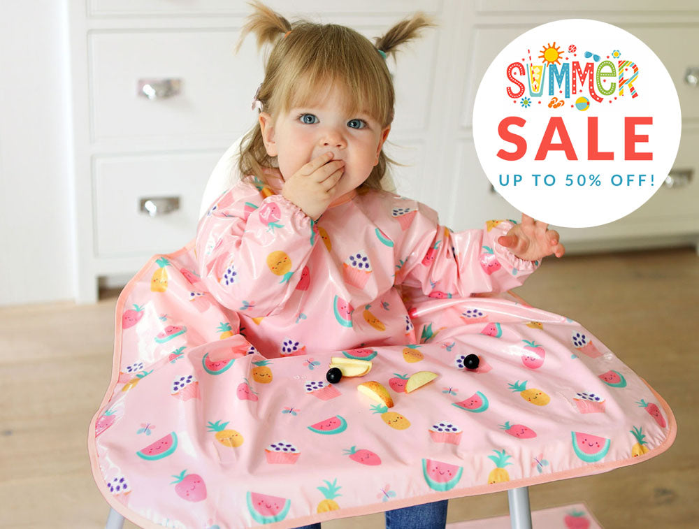A happy toddler child sat in a highchair wearing a Bibetta Wipeezee pink Yummy Treats fruit and cakes print coverall baby bib that covers the highchair table with a 'Summer Sale' graphic in the corner.