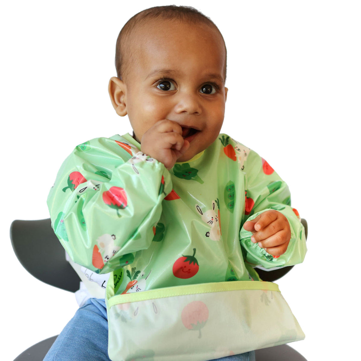 a smiling toddler seated in a highchair wearing a Bibetta wipeezee sleeved bib in green bunny and veg pattern with the pocket folded to the front. The baby is eating a piece of food and his hand is in front of his mouth.