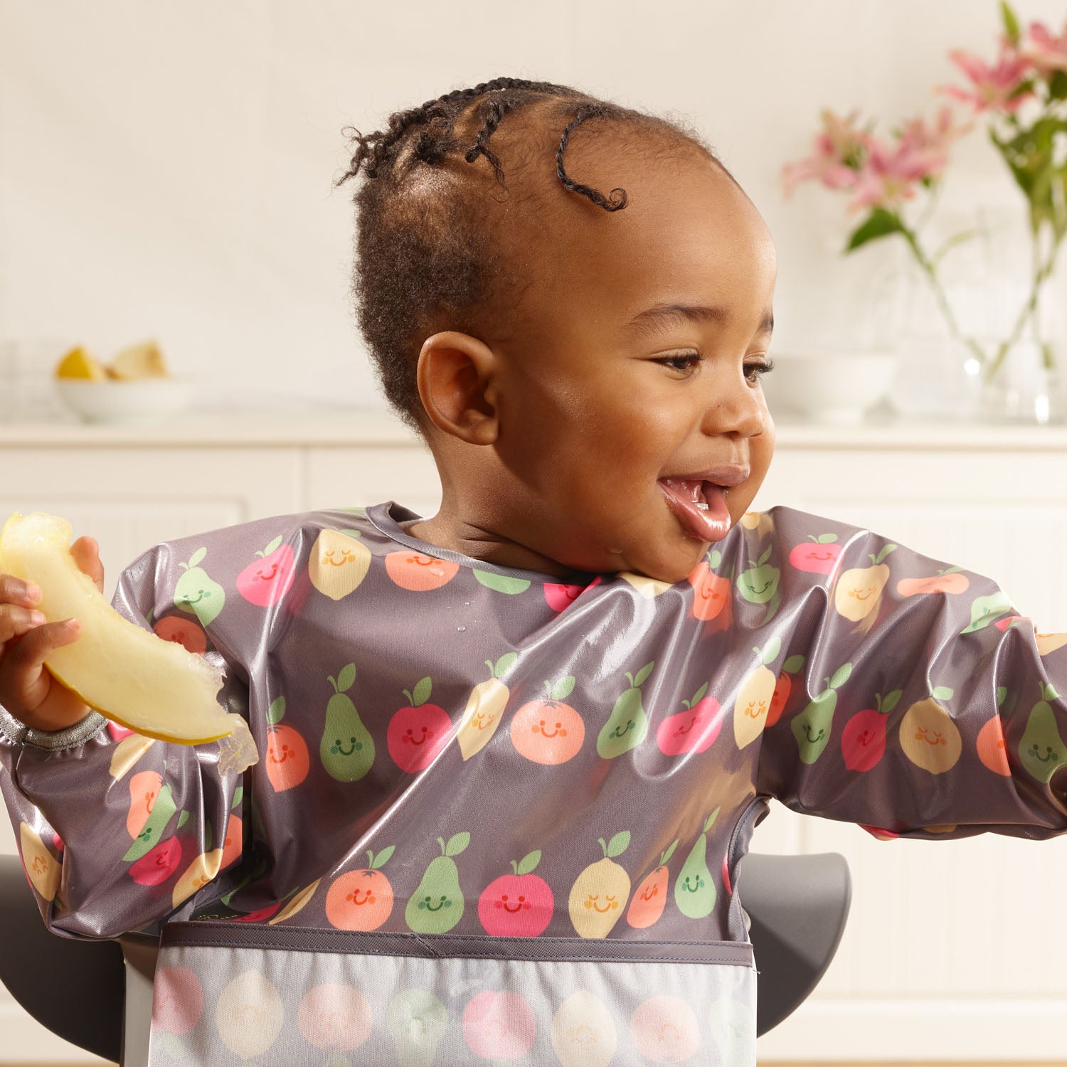 a smiling toddler seated in a highchair wearing a Bibetta wipeezee sleeved bib in grey happy fruit pattern with the pocket folded to the front. The baby is eating a piece of melon.