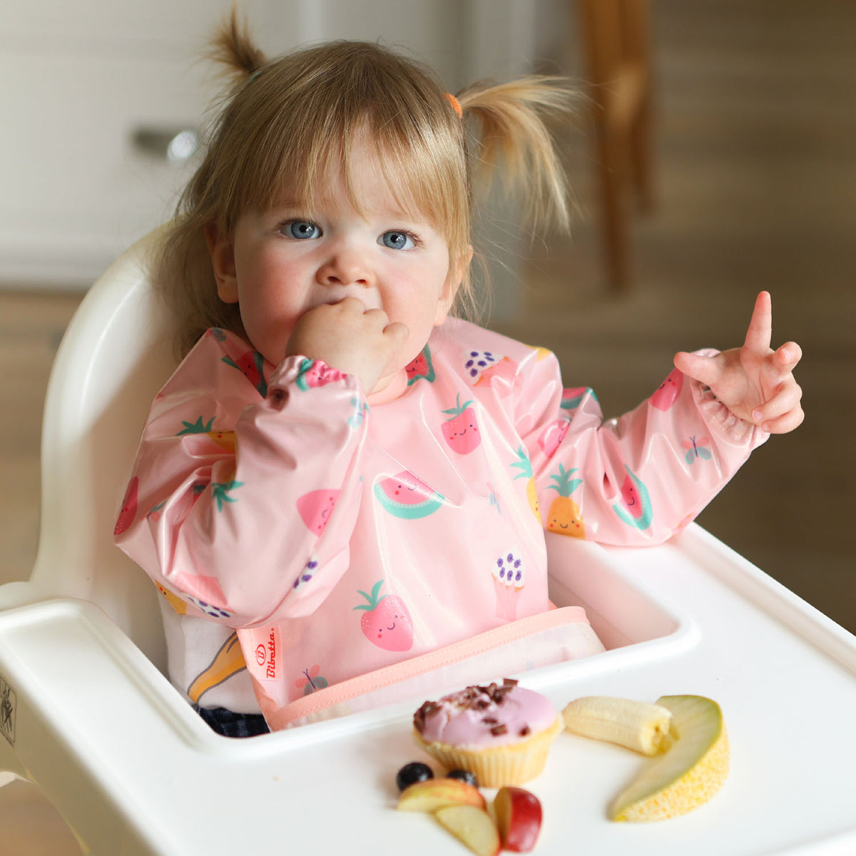 a toddler seated in a highchair wearing a Bibetta wipeezee sleeved bib in pink yummy treats pattern with the pocket folded to the front. The baby is eating a piece of food from the selection on the highchair tray.