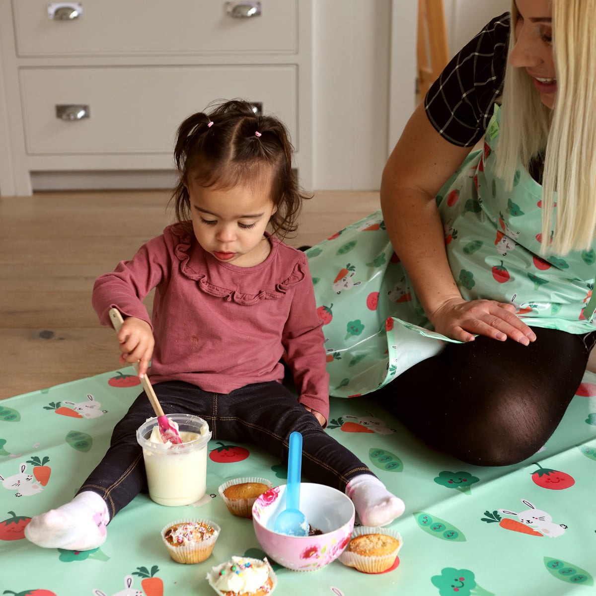 a little girl and her mum sitting on a Wipeezee splash mat in green bunny and veg on the floor. The littel girl sis weaing a pink top and denim leggings. She is playing with icing in a jar with a spatula. Her mum is watching and is wearing a black dress and tights and a Wipeezee adult apron in green bunny and veg.