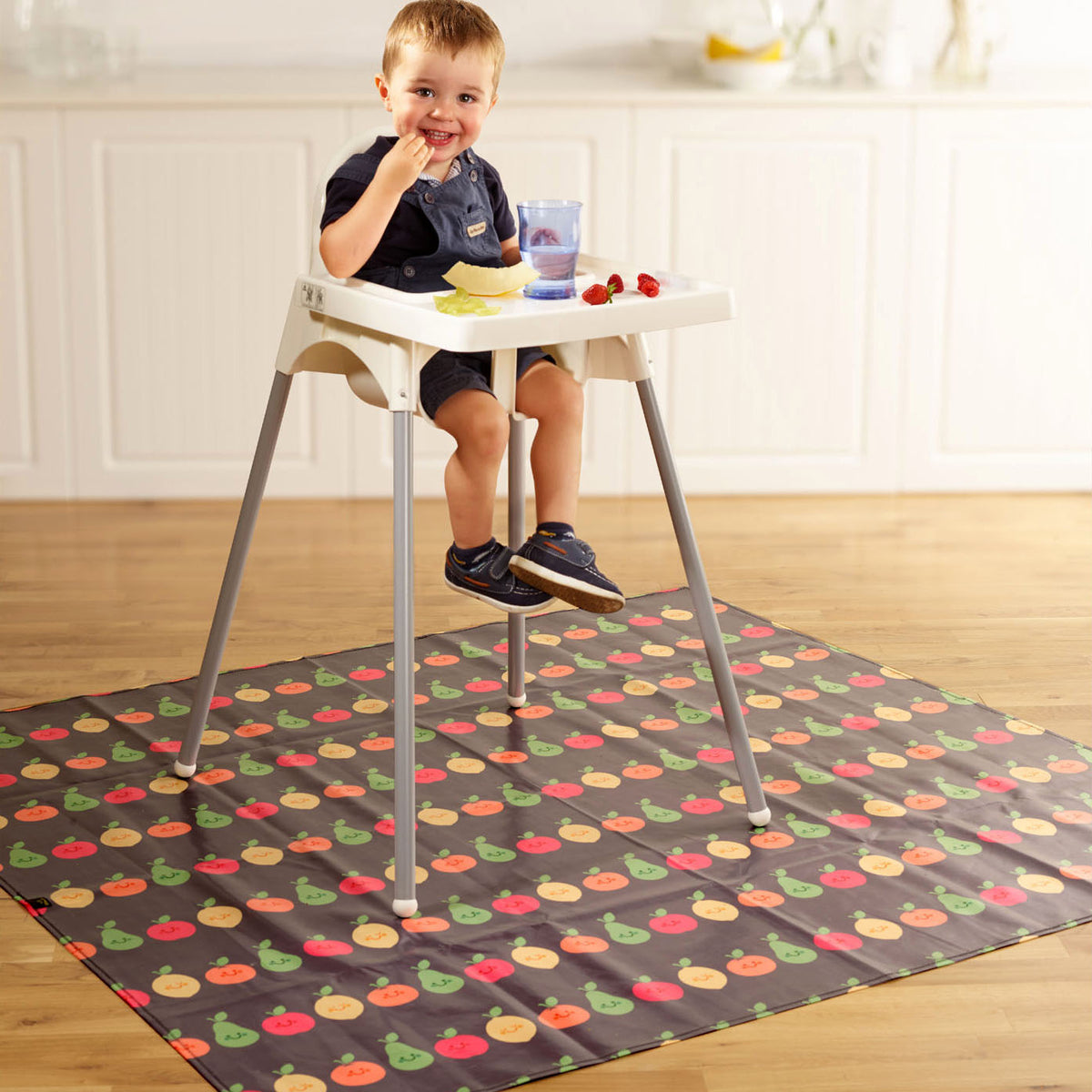 little boy seated in a highchair eating melon and strawberries. Underneath and on the floor is a wipeezee splash mat in grey happy fruits