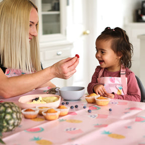 little girl sat at the table with her mum playing with cakes and fruit. The table has a wipeezee splash mat in pink yummy treats on it. Both mum and daughter are wearing matching wipeezee aprons in the same pink colour.