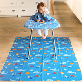 little boy seated in a highchair eating spaghetti and wearing a wipeezee turquoise sea creatures coverall bib which is stretched over the highchair tray. Underneath and on the floor is a matching wipeezee splash mat in the same colour.