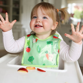 a smiling toddler seated in a white ikea highchair wearing a Bibetta wipeezee weaning bib in green bunny and veg pattern with the pocket folded to the front. The baby is dressed in a white long sleeved top and has her hands in the air.