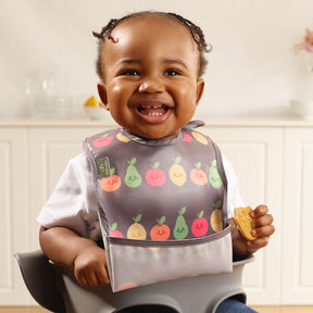 a smiling baby seated in a grey highchair wearing a Bibetta wipeezee weaning bib in grey happy fruits pattern with the pocket folded to the front. The baby is dressed in a white t-shirt and denim jeans. 