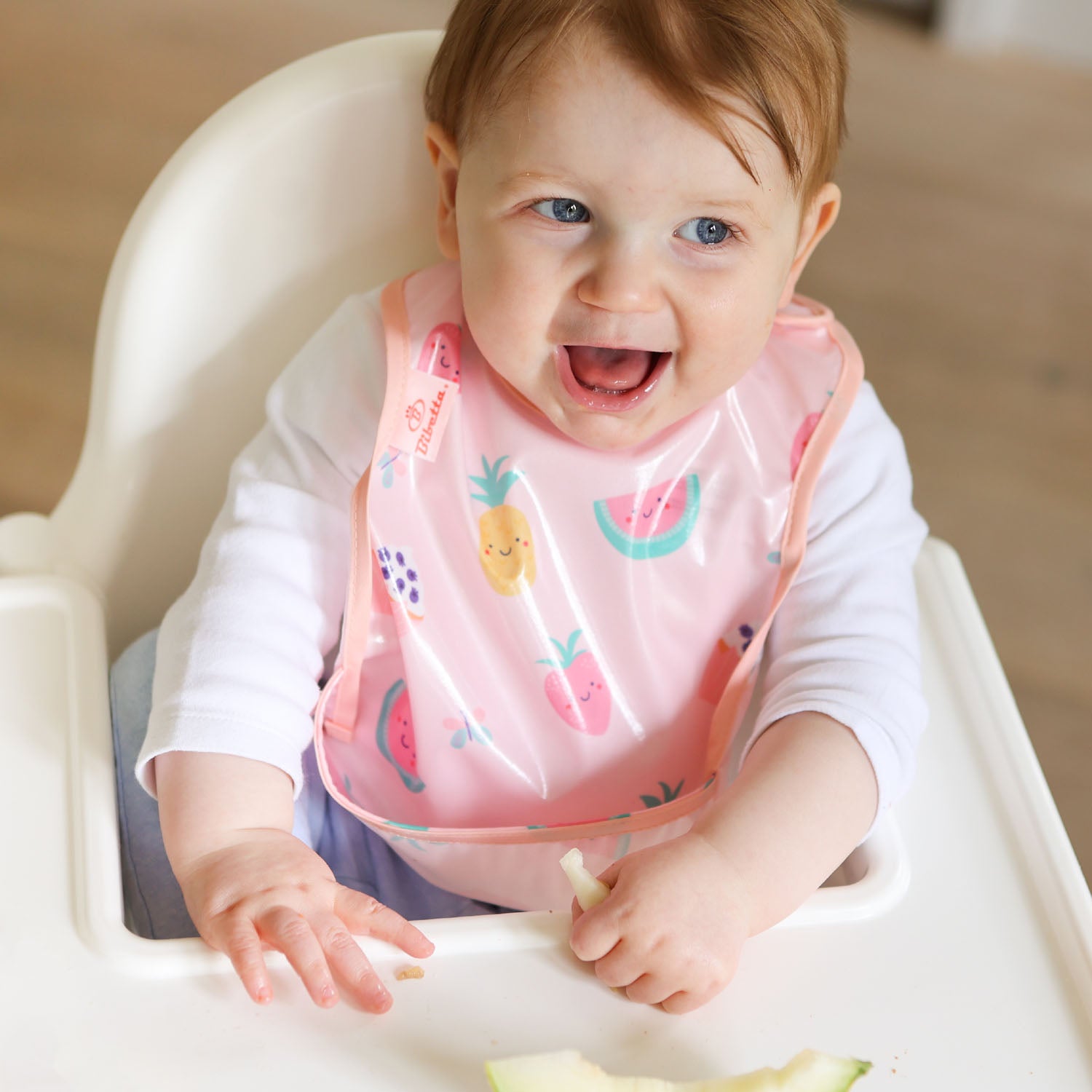 a smiling baby seated in a white ikea highchair wearing a Bibetta wipeezee weaning bib in pink yummy treats pattern with the pocket folded to the front. The baby is dressed in a white long sleeved top and blue leggings. 