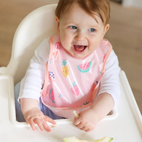 a smiling baby seated in a white ikea highchair wearing a Bibetta wipeezee weaning bib in pink yummy treats pattern with the pocket folded to the front. The baby is dressed in a white long sleeved top and blue leggings. 