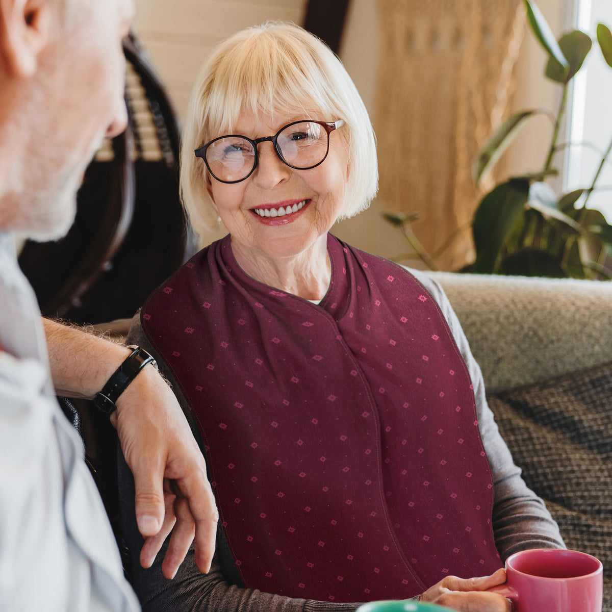 Woman with a blonde bob haircut, dark brown glasses wearing a brown jumper and a burgundy straight scarf clothing protector sitting next to a man, holding a mug.