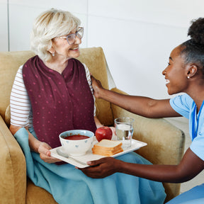 Woman serving a tray of food to an elderly woman on a couch who is wearing a burgundy straight scarf clothing protector 