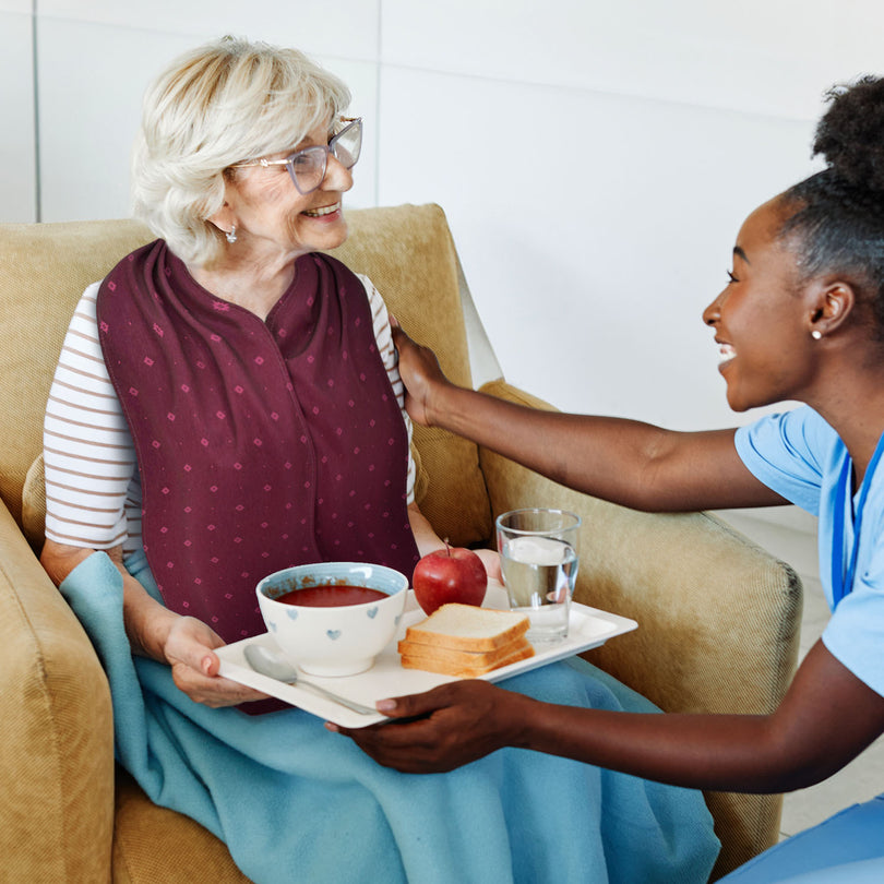 Woman serving a tray of food to an elderly woman on a couch who is wearing a burgundy straight scarf clothing protector 