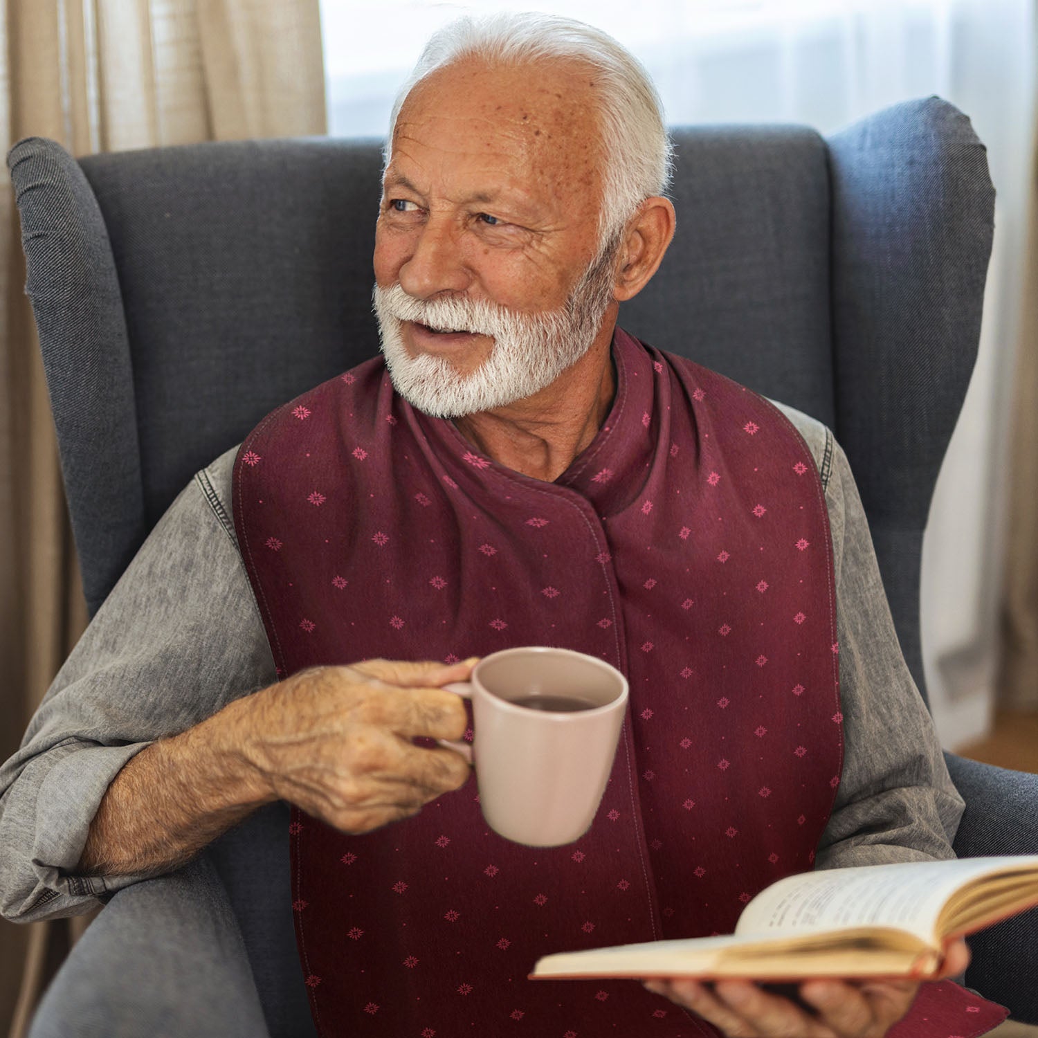 Older gentleman sitting in a chair holding a cup and book, wearing a burgundy patterned straight scarf clothing protector 