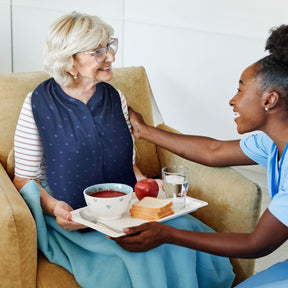 Woman serving a tray of food to an elderly woman on a couch who is wearing a navy blue straight scarf clothing protector 