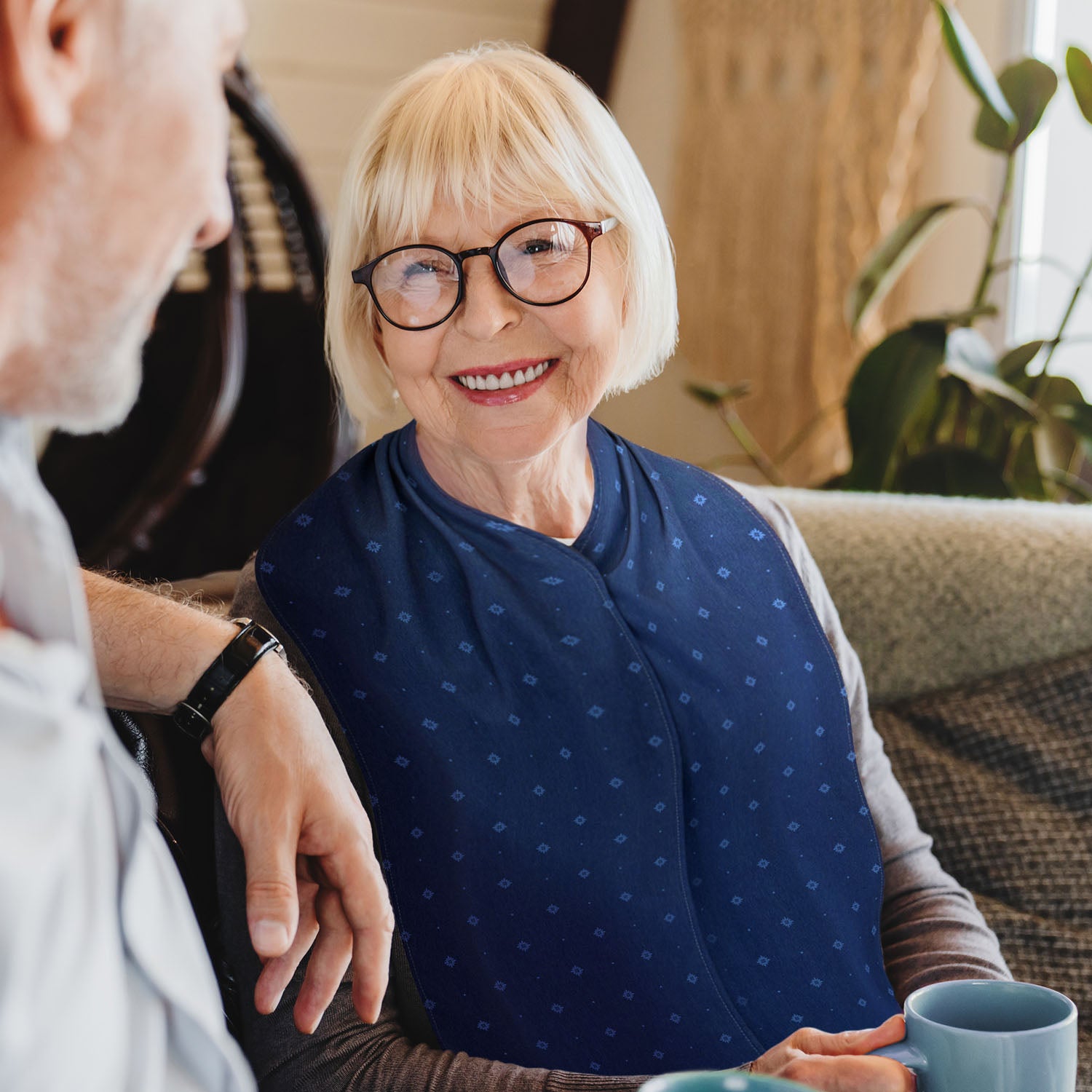 Woman with a blonde bob haircut, dark brown glasses wearing a brown jumper and navy blue straight scarf clothing protector sitting next to a man, holding a mug.