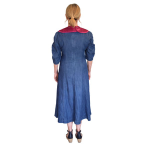 A woman viewed from the back wearing a long blue dress and a crae designs tabard bib and dark wedge sandals, set against a plain white background.