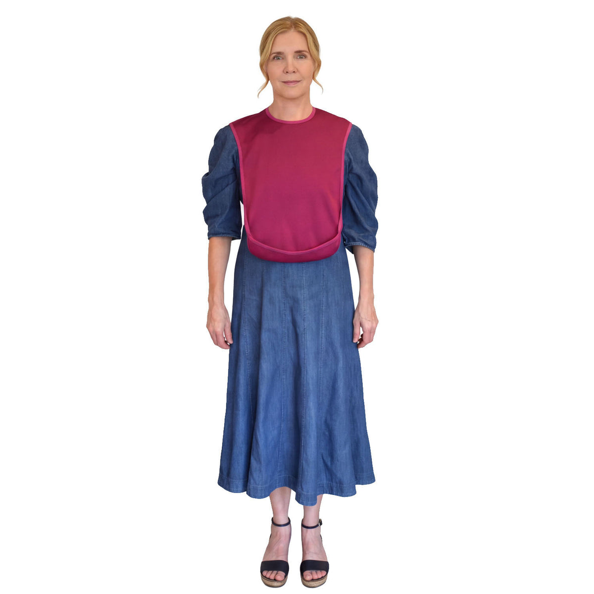 Lady standing wearing a blue dress and a Carea Designs Small burgundy tabard bib with the pocket facing out to the front. 