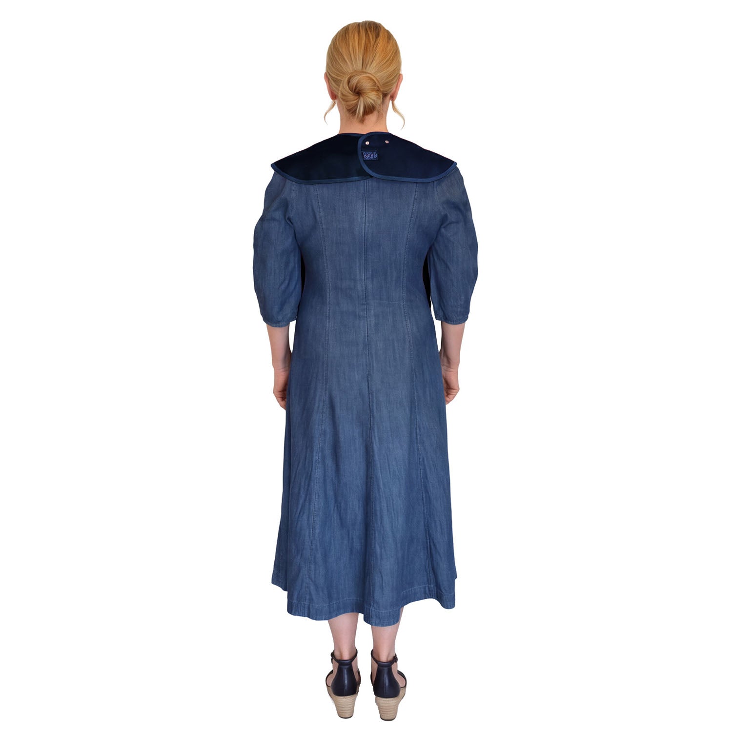A woman viewed from the back wearing a blue midi dress with a large navy Care Designs tabard adult bib and dark navy wedge sandals, set against a plain white background.