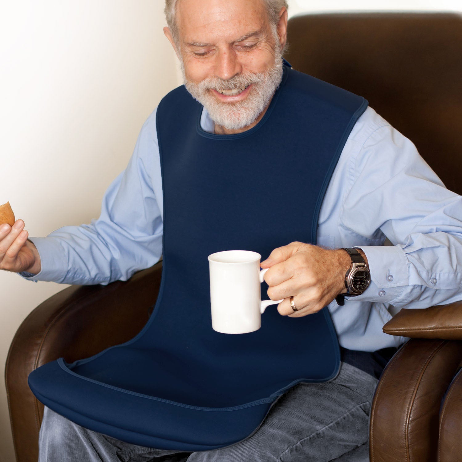 A man wearing a Care Designs large navy blue tabard adult bib sits in a brown leather armchair, holding a white mug. The bib has a wide curved bottom tray and is worn over a light blue button-down shirt.