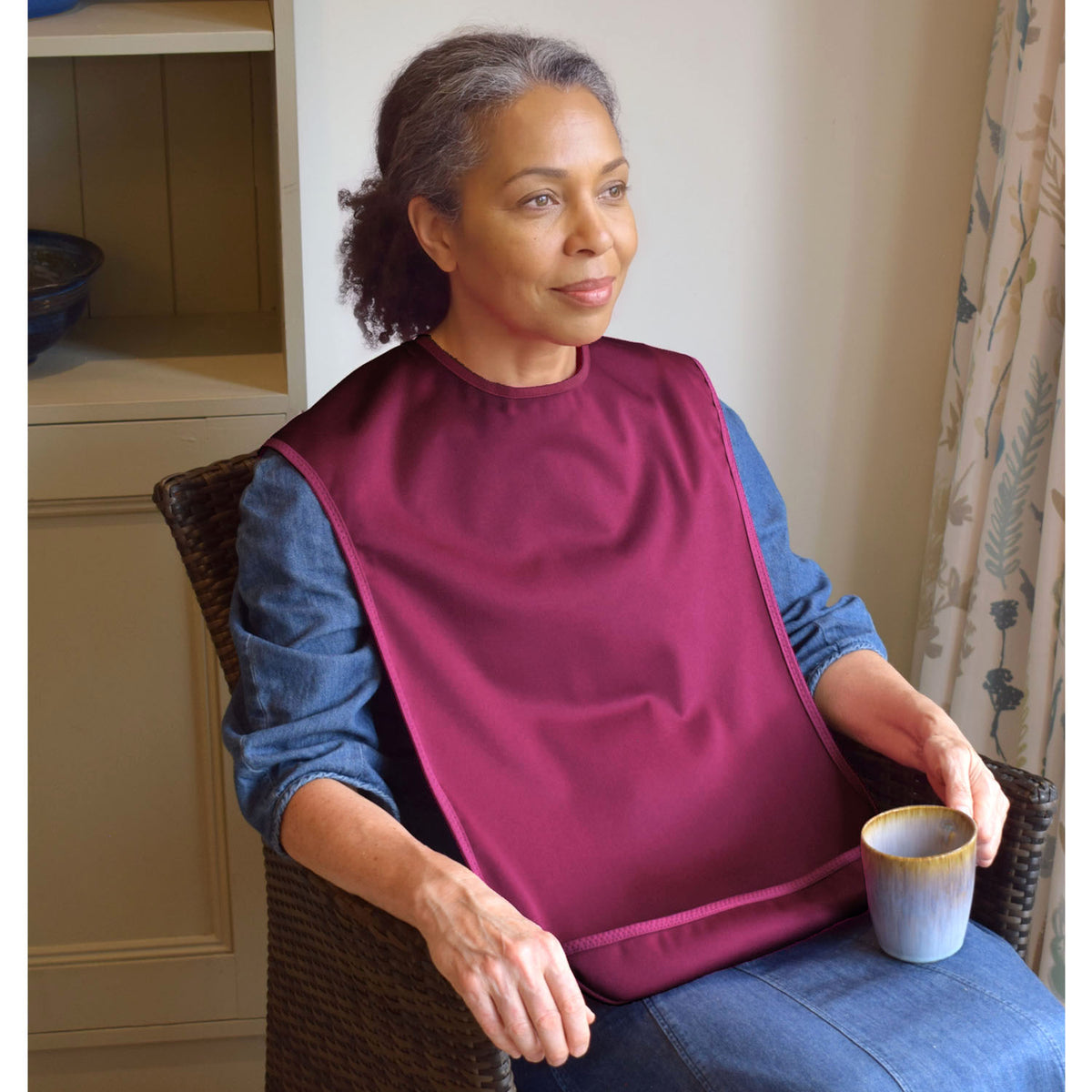 A woman seated in a wicker chair indoors wearing a Care Designs burgundy medium tabard style bib  over a blue long-sleeve top, holding a small cup.