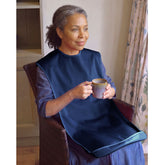 A woman sitting in a brown armchair indoors, wearing a Care Designs large navy tabard adult bib and holding a small ceramic mug.