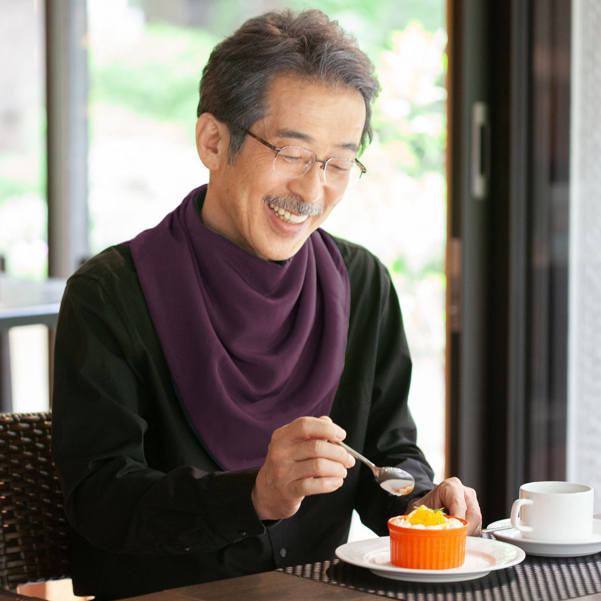 Man eating dessert with a spoon at a table, wearing glasses and an aubergine long neckerchief bib.
