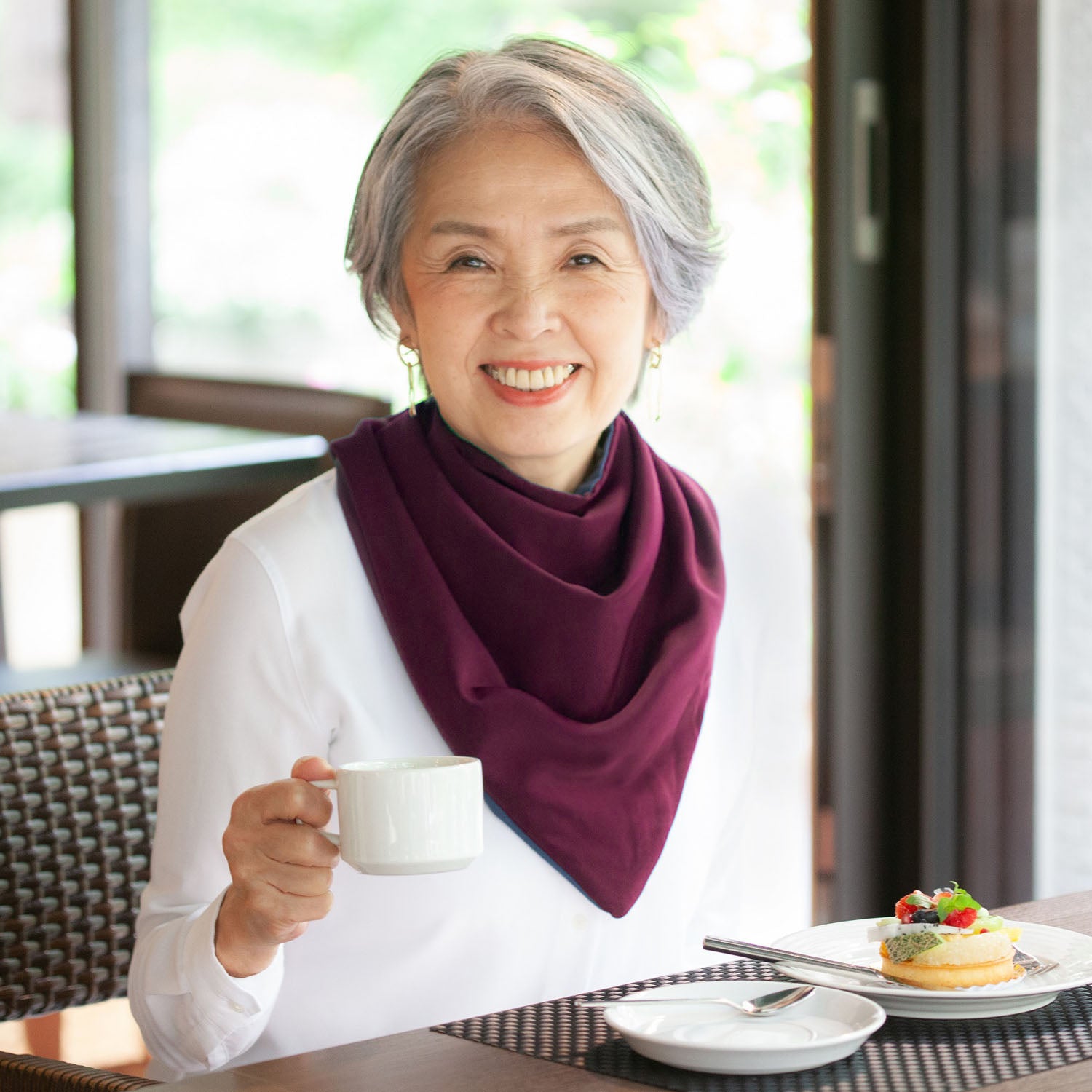 Woman sitting at a table with a cup of coffee and dessert, wearing a burgundy long neckerchief bib.