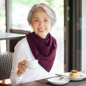 Woman sitting at a table with a cup of coffee and dessert, wearing a burgundy long neckerchief bib.