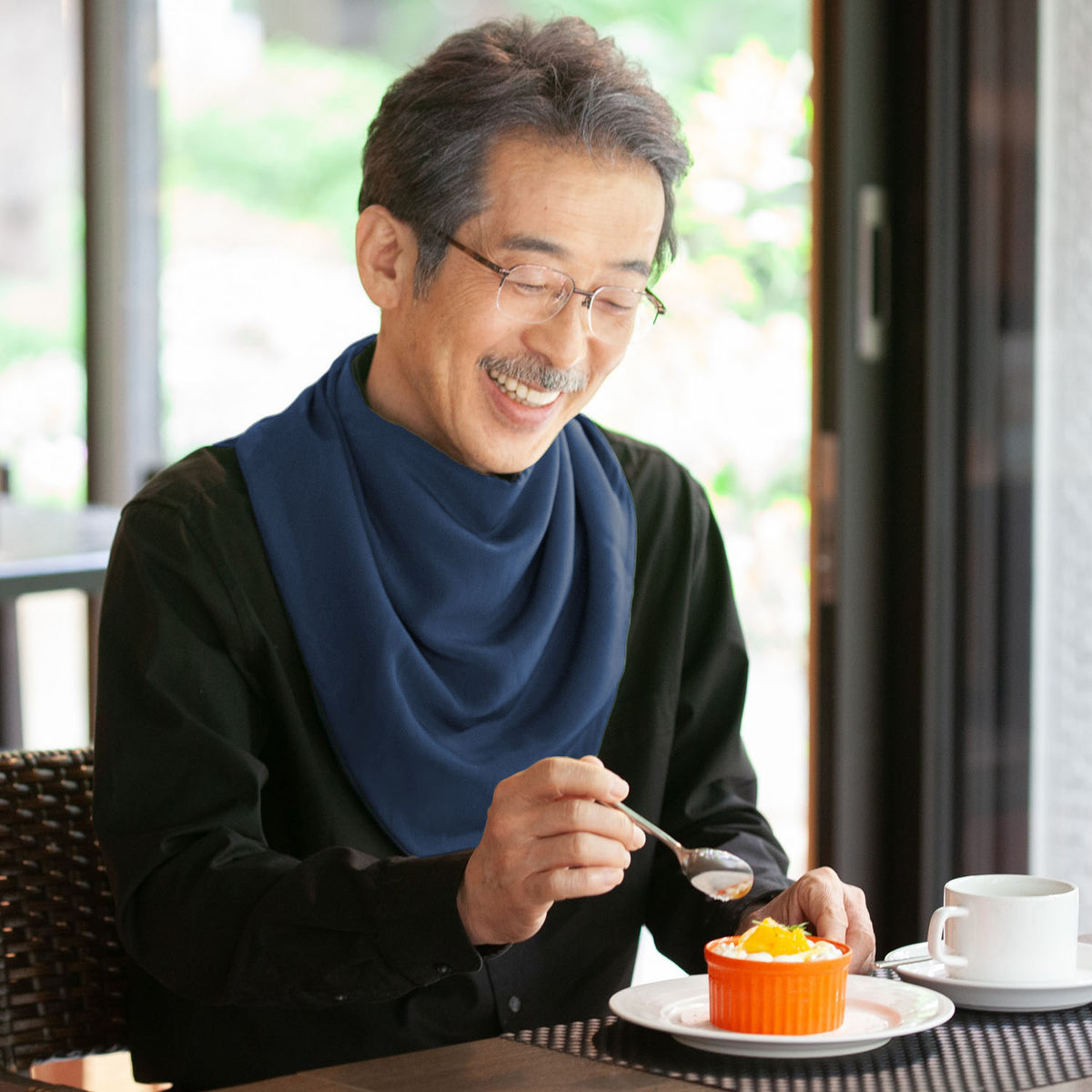 Man wearing a long navy neckerchief bib enjoying dessert at a cafe with a blue scarf and glasses.