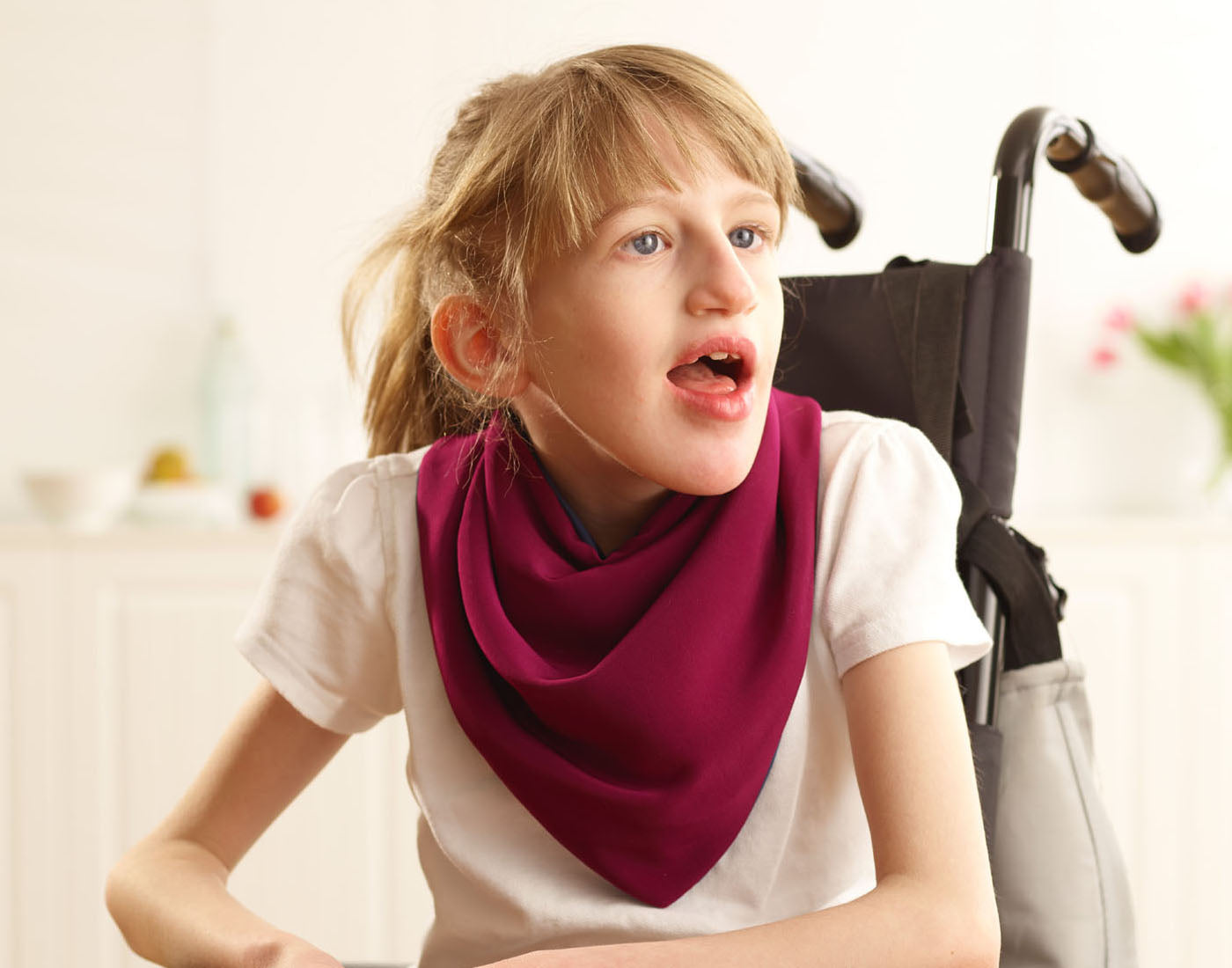 Young girl sitting in a wheelchair wearing a Care Designs burgundy neckerchief style adult bib clothing and dribble protector
