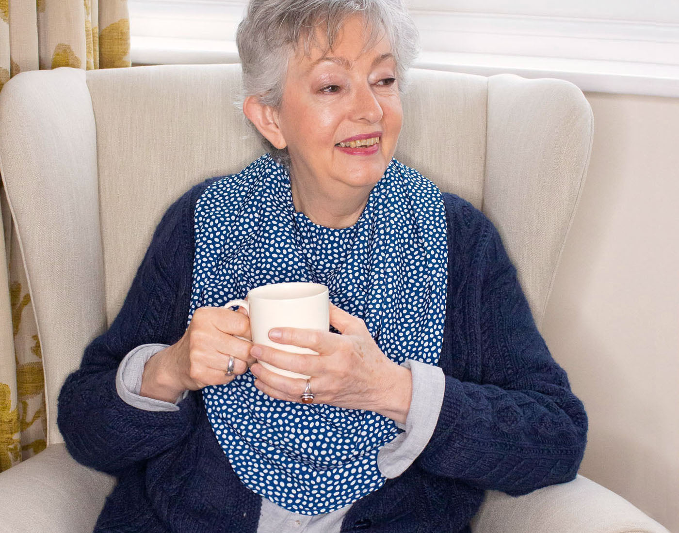 Senior woman sitting in a chair wearing a Care Designs Navy dot pashmina scarf style adult bib clothing protector drinking a cup of tea.
