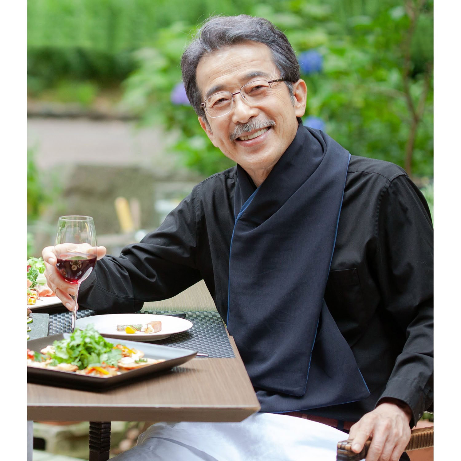 Man sitting at an outdoor table  wearing a black cross scarf clothing protector with a glass of red wine and a plate of food, surrounded by greenery.