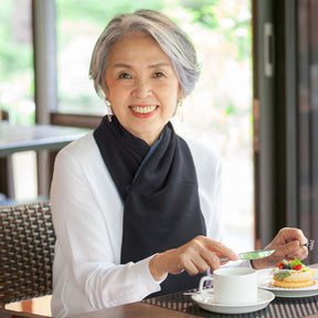 Woman sitting at a table wearing a white top and a black cross scarf clothing protector with a cup of tea and a plate of food, smiling.
