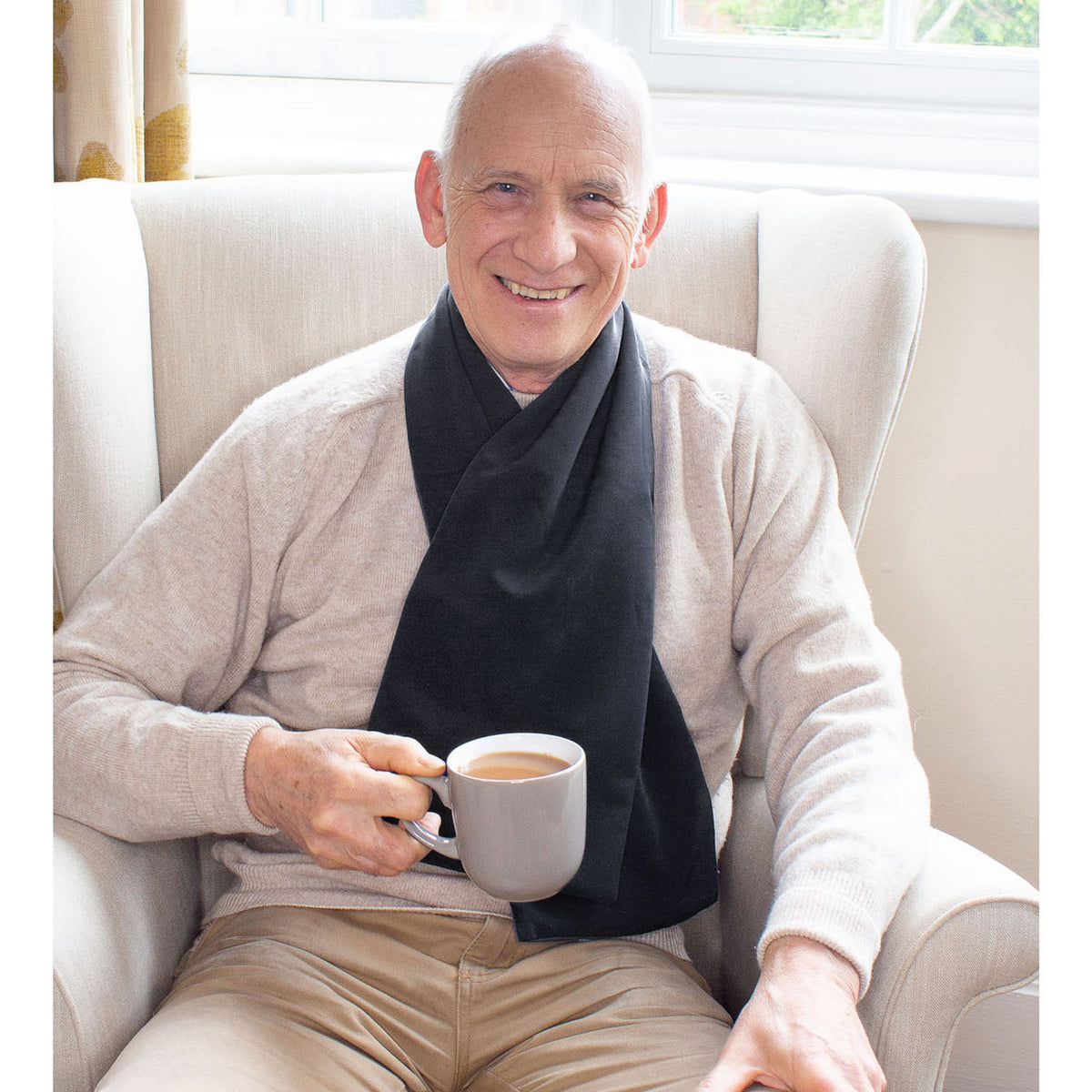 Man sitting in a chair holding a cup of coffee, wearing a beige sweater and black cross scarf clothing protector.
