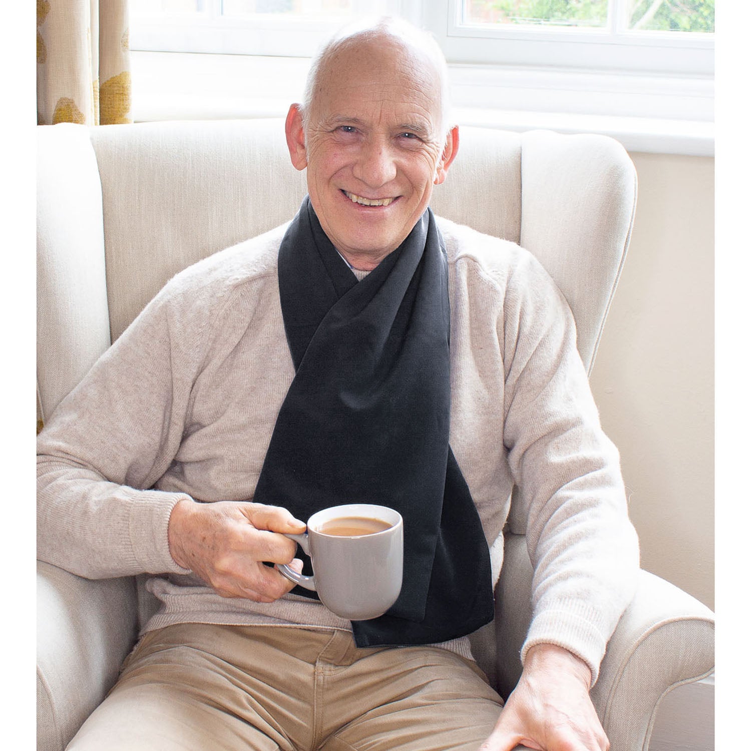 Man sitting in a chair holding a cup of coffee, wearing a beige sweater and black cross scarf clothing protector.