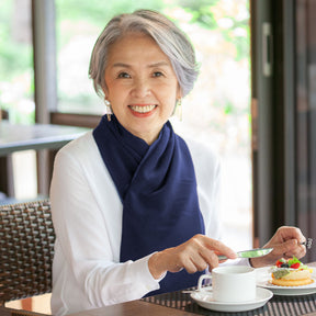 Woman sitting at a table with a cup of tea and a plate of food, wearing a navy blue cross scarf clothing protector.