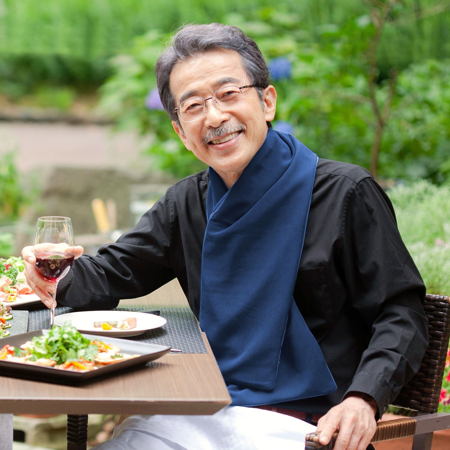 Man sitting at an outdoor table with a glass of red wine and a plate of food, surrounded by greenery wearing a navy cross scarf clothing protector.
