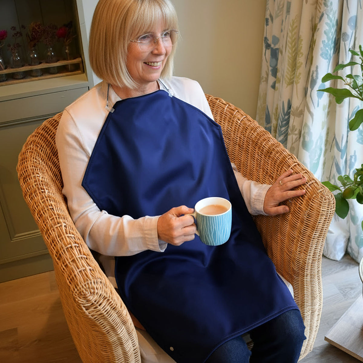 Lady sitting in a chair holding a cup of tea in her right hand.
She is wearing a white shirt and blue jeans and has a a Care Designs long waterproof navy napkin tucked into her shirt collar and held up with delicate straps arounf her neck.