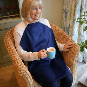 Lady sitting in a chair holding a cup of tea in her right hand.
She is wearing a white shirt and blue jeans and has a a Care Designs long waterproof navy napkin tucked into her shirt collar and held up with delicate straps arounf her neck.