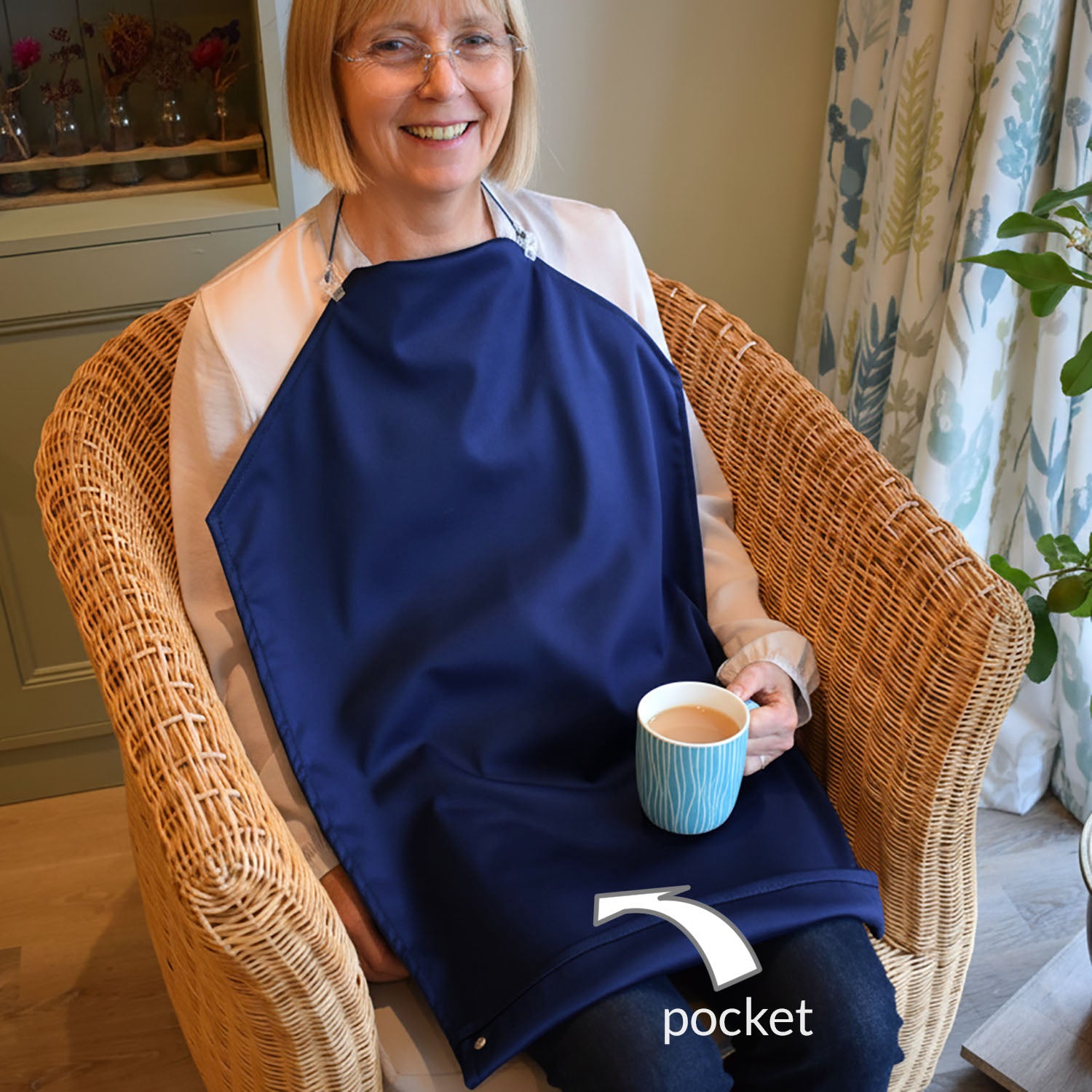 Lady sitting in a chair holding a cup of tea in her left hand.
She is wearing a white shirt and blue jeans and has a a Care Designs long waterproof navy napkin tucked into her shirt collar and held up with delicate straps arounf her neck. The napkin is folded over at the bottom and held up with press studs.