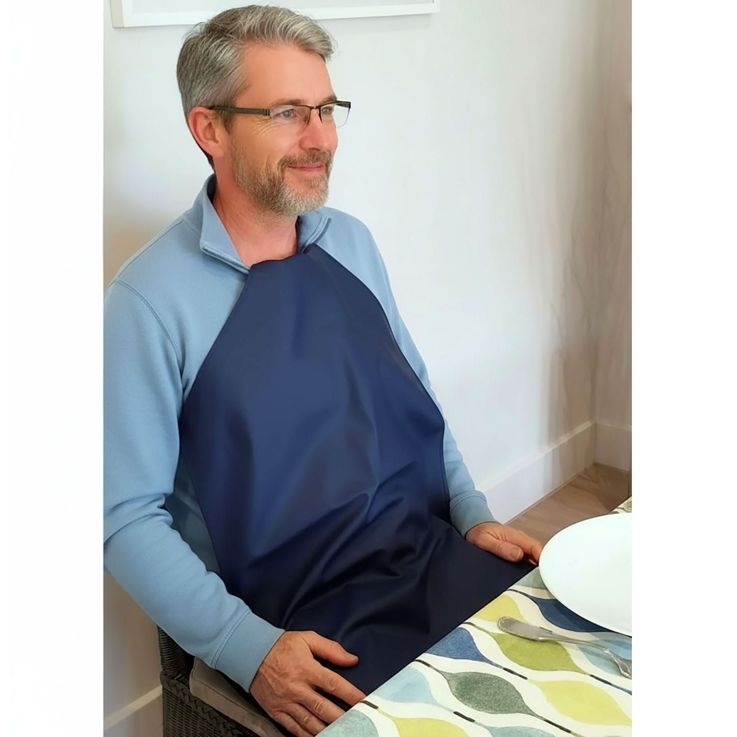 Man wearing a Care Designs long navy waterproof napkin tucked into his collar sitting at a table with a white wall background