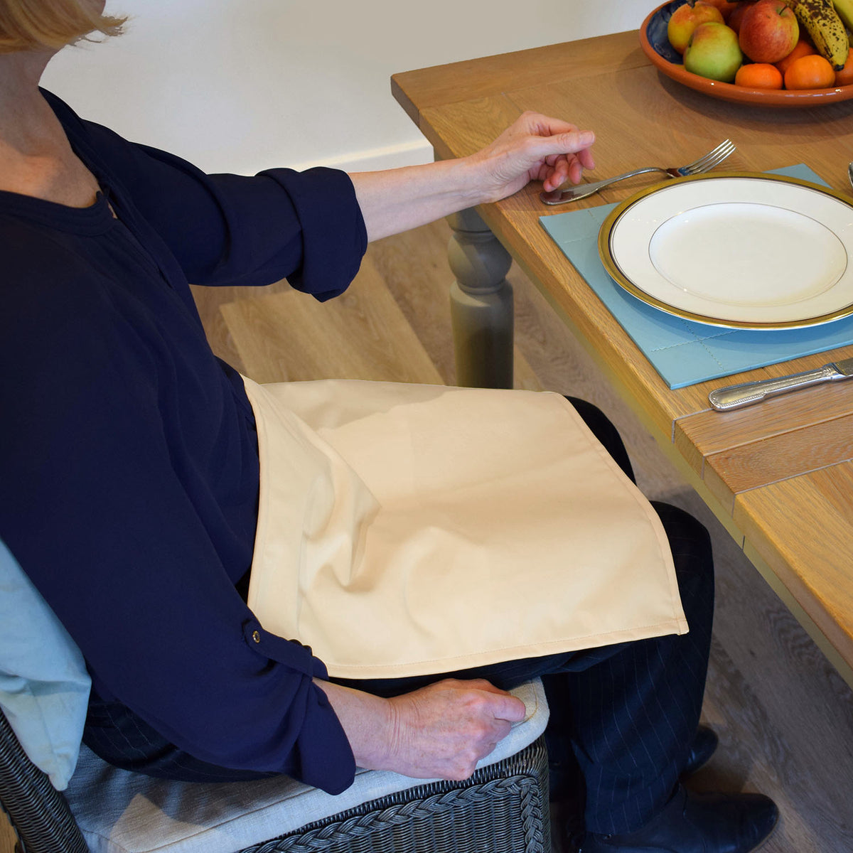 Person sitting at a table with a plate and fork, wearing a blue shirt and navy trousers with a Care Designs cream waterproof napkin on their lap.