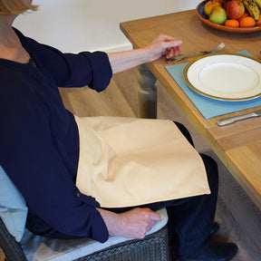 Person sitting at a table with a plate and fork, wearing a blue shirt and navy trousers with a Care Designs cream waterproof napkin on their lap.